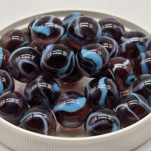 May include: A collection of glass marbles in a white dish. The marbles are a mix of dark red, black, and light blue, with swirling patterns. The marbles are round and have a glossy finish.
