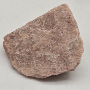 May include: A rough, irregularly shaped rock with a muted pinkish-brown hue. The surface has a textured appearance with subtle variations in color and tone. The rock is set against a plain white background.