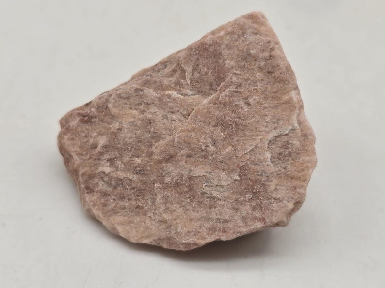 May include: A rough, irregularly shaped rock with a muted pinkish-brown hue. The surface has a textured appearance with subtle variations in color and tone. The rock is set against a plain white background.