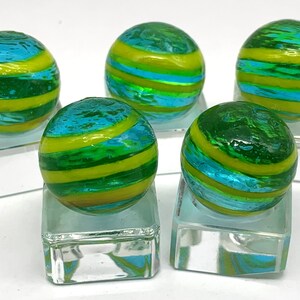 K&ouml;nnte beinhalten: F&uuml;nf Glasmurmeln mit einem Wirbeldesign. Die Murmeln sind gr&uuml;n, blau und gelb und werden auf transparenten Glasst&auml;ndern pr&auml;sentiert. Die Murmeln haben eine gl&auml;nzende Oberfl&auml;che und sind rund.