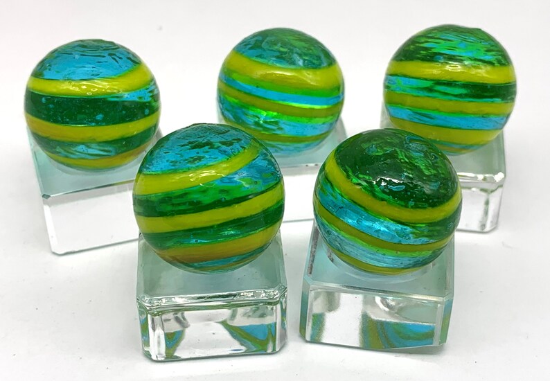 K&ouml;nnte beinhalten: F&uuml;nf Glasmurmeln mit einem Wirbeldesign. Die Murmeln sind gr&uuml;n, blau und gelb und werden auf transparenten Glasst&auml;ndern pr&auml;sentiert. Die Murmeln haben eine gl&auml;nzende Oberfl&auml;che und sind rund.