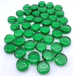 May include: A large group of green glass pebbles, also known as marbles or gems, are scattered on a white surface.
