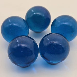 May include: Five translucent blue glass marbles arranged in a circular pattern. The marbles have a glossy finish and varying shades of blue, from light to deep. They are set against a plain white background.