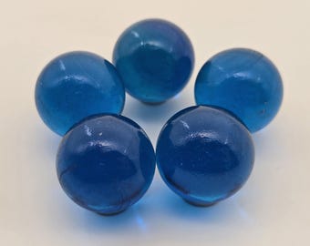May include: Five translucent blue glass marbles arranged in a circular pattern. The marbles have a glossy finish and varying shades of blue, from light to deep. They are set against a plain white background.