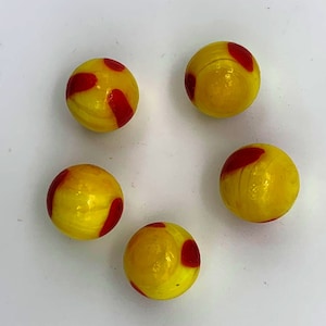 May include: Five yellow glass marbles with red swirls. The marbles are arranged in a circle on a white surface.