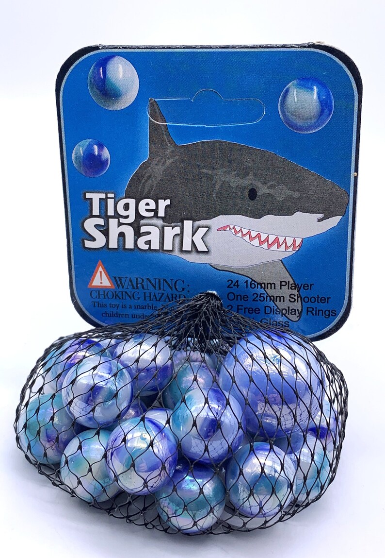 Puede incluir: Una bolsa de 24 canicas de vidrio azul y blanco con una ilustraci&oacute;n de tibur&oacute;n tigre en el empaque. El empaque tambi&eacute;n incluye el texto "Tiger Shark", "WARNING: CHOKING HAZARD", "This toy is a marble", "children under", "24 16mm Player", "One 25mm Shooter", "2 Free Display Rings", y "Glass".
