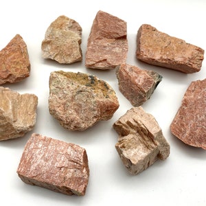 May include: A collection of rough, natural stones in shades of pink, beige, and reddish-brown. The stones vary in size and shape, with visible textures and mineral formations. They are arranged on a white surface, showcasing their natural colors and details.
