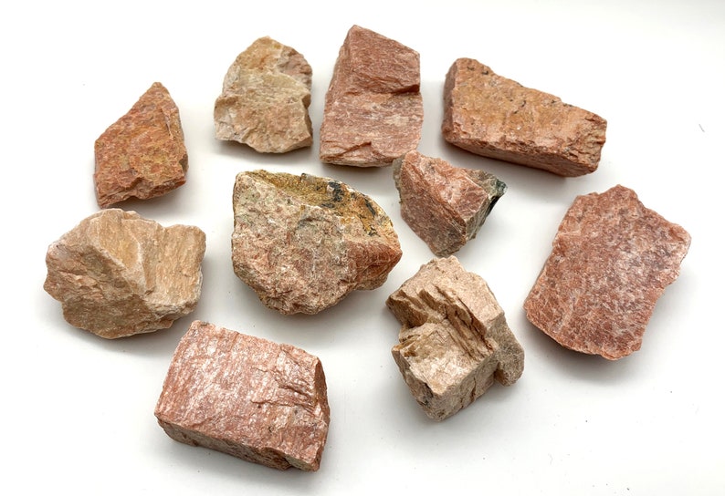 May include: A collection of rough, natural stones in shades of pink, beige, and reddish-brown. The stones vary in size and shape, with visible textures and mineral formations. They are arranged on a white surface, showcasing their natural colors and details.