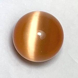 May include: A round, polished gemstone with a warm, peach-colored hue. The stone has a prominent, light-colored band across its center, creating a cat's eye effect. The surface is smooth and reflective, with a subtle sheen.