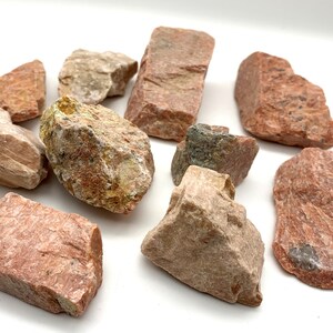 May include: A collection of rough-hewn rocks in shades of red, tan, and gray. The stones vary in size and shape, with visible textures and mineral patterns. The rocks are scattered on a white surface, showcasing their natural colors and forms.