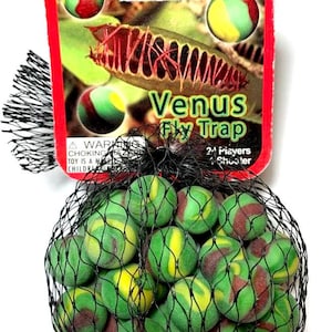 May include: A Venus Fly Trap game set. The set includes green, yellow, and maroon marbles in a black mesh bag. The packaging features a red border with the game's name and a graphic of a Venus flytrap.
