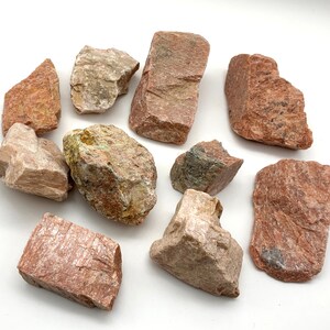 May include: A collection of rough-cut rocks in shades of red, tan, and gray, scattered on a white surface. The stones vary in size and shape, with visible textures and natural variations. The image showcases a variety of geological specimens.