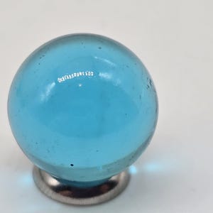 May include: A clear, light blue glass marble with a diameter of approximately 1 inch, mounted on a small, silver-colored metal base. The marble is spherical and translucent, with slight imperfections.