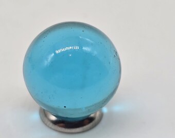 May include: A clear, light blue glass marble with a diameter of approximately 1 inch, mounted on a small, silver-colored metal base. The marble is spherical and translucent, with slight imperfections.