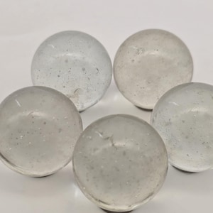 May include: Five clear glass spheres with internal bubbles are arranged against a white background. The spheres are translucent, allowing light to pass through, and have a smooth, rounded shape. They appear to be decorative or collectible items.