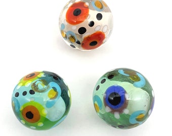 May include: Three glass marbles with colorful abstract patterns. The marbles are white, green, and blue with red, orange, yellow, and black accents.