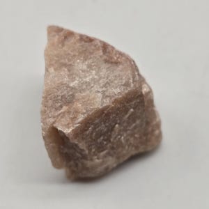 May include: A rough, uncut gemstone with a light brown color. The stone has a textured surface and irregular shape, with visible crystalline structures. The gemstone is set against a plain white background.