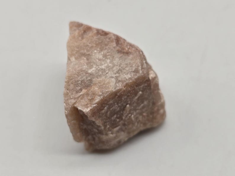 May include: A rough, uncut gemstone with a light brown color. The stone has a textured surface and irregular shape, with visible crystalline structures. The gemstone is set against a plain white background.