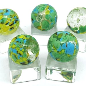 May include: Five glass marbles with green, blue, and yellow swirls on a white background. The marbles are displayed on clear square bases.