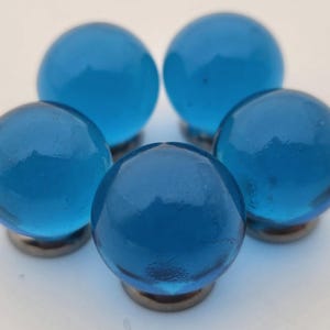 May include: Five round, transparent blue glass cabinet knobs with a faceted surface. Each knob is mounted on a small, silver-colored metal base. The knobs are arranged in a circular pattern against a white background.