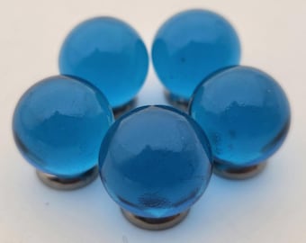 May include: Five round, transparent blue glass cabinet knobs with a faceted surface. Each knob is mounted on a small, silver-colored metal base. The knobs are arranged in a circular pattern against a white background.