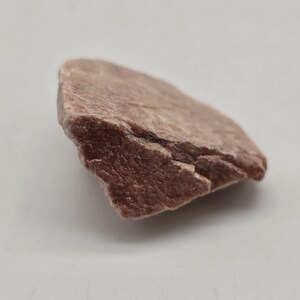May include: A single, irregularly shaped piece of raw, reddish-brown stone. The stone has a rough, textured surface with visible cracks and variations in color. The background is a plain, light gray.