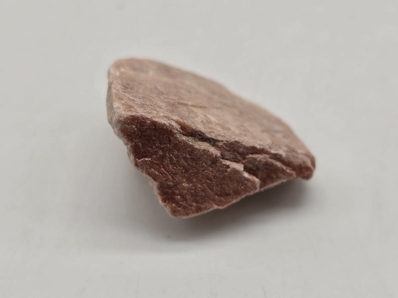 May include: A single, irregularly shaped piece of raw, reddish-brown stone. The stone has a rough, textured surface with visible cracks and variations in color. The background is a plain, light gray.
