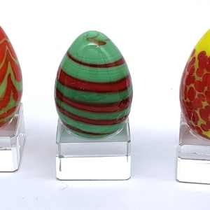 May include: Three decorative glass egg sculptures. The eggs are displayed on clear acrylic stands. One egg is red with green swirls, another is green with red stripes, and the third is yellow with red spots.
