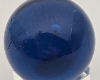 May include: A deep blue glass marble with a glossy finish. The marble is spherical and appears to have small air bubbles within the glass. It is displayed on a clear stand, showcasing its color and shape.