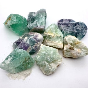 May include: A collection of seven green, purple, and white fluorite crystals. The crystals are rough and have a variety of shapes and sizes.