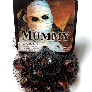 May include: A bag of 24 brown and black marbles with a white swirl design. The bag has a warning label that says "WARNING: CHOKING HAZARD TOY IS A MARBLE. NOT FOR CHILDREN UNDER 3 YRS". The label also says "24 Players 1 Shooter". The bag is in front of a card with a mummy on it. The card says "MUMMY" in large letters.