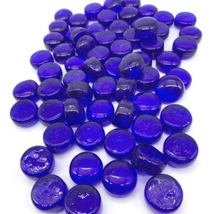 May include: A large group of cobalt blue glass pebbles. The pebbles are smooth and round, and they are scattered on a white surface.