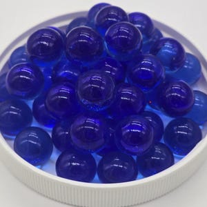 May include: A collection of deep blue glass marbles fills a white, round container. The marbles are smooth and reflective, with a glossy finish. The container has a ribbed texture on the side. The marbles are a vibrant, saturated color.