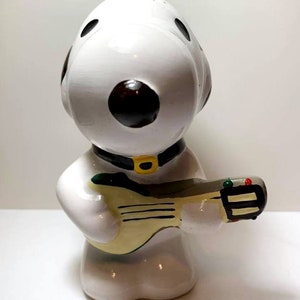 May include: A white ceramic figurine of a cartoon dog with black spots and a black collar with a yellow buckle. The dog is holding a light yellow guitar with green and red accents.