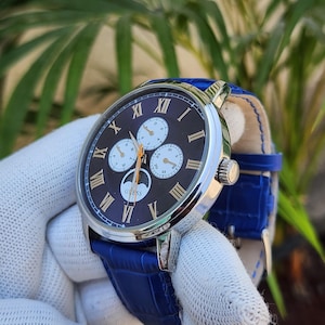 May include: A silver-tone analogue wristwatch with a dark brown dial, gold-coloured Roman numeral hour markers, and three sub-dials. The watch features a moon phase indicator above the 6 o'clock position with the brand name Guess printed in white.