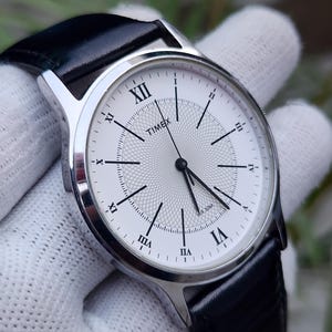 Könnte beinhalten: Eine Timex Armbanduhr mit silberfarbenem, rundem Gehäuse, weißem Zifferblatt mit zentralem Guilloché-Muster, schwarzen römischen Ziffern und dünnen schwarzen Zeigern. Die Uhr hat ein schwarzes Lederarmband und die Aufschrift WR 30M.