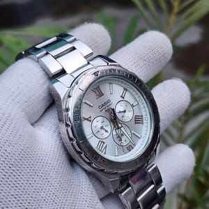 Pode incluir: Um relógio de pulso Casio prateado com mostrador branco, apresentando algarismos romanos, três submostradores e uma pulseira de elos de aço inoxidável. O mostrador exibe claramente a marca Casio e o texto Water Resist 100M.