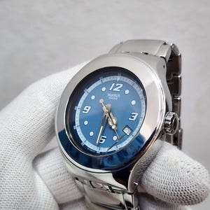 Puede incluir: Un reloj de pulsera Swatch de acero inoxidable en tono plateado con una esfera redonda azul, números arábigos blancos en las 12, 3, 6 y 9, una ventana de fecha en la posición de las 4, manecillas luminosas y una correa de eslabones metálicos.