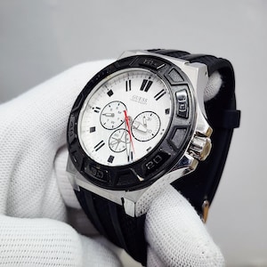 Op de afbeelding: Een Guess polshorloge met een witte wijzerplaat, drie sub-wijzerplaten en een zwarte, gestructureerde siliconen band. Het horloge heeft een zilverkleurige roestvrijstalen kast, een zwarte draaibare ring met cijfers en een rode secondewijzer.