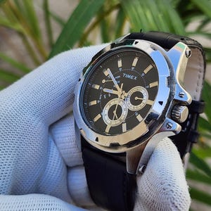 Pode incluir: Um relógio de pulso Timex com caixa redonda de metal prateado e mostrador preto com ponteiros e marcadores luminosos. O relógio inclui três submostradores para o dia e a data, uma pulseira de couro preto texturizado e uma coroa de ajuste lateral.