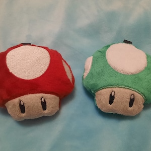 Op de afbeelding: Twee pluche paddenstoel speelgoed, een rood en een groen, met witte stippen en grijze gezichten.