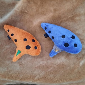 May include: Two plush ocarinas, one orange with black dots and one blue with black dots. Both ocarinas have a green triangle on the bottom.
