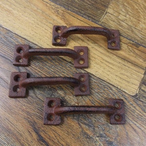 May include: Four rustic brown cast iron drawer pulls with square ends and a curved handle. The pulls are arranged in a row on a wooden surface.