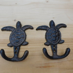 May include: Two black cast iron turtle hooks with double hooks. The hooks are mounted on a wooden surface.