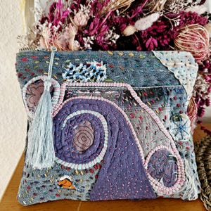 Könnte beinhalten: Eine Denim-Patchwork-Clutch mit aufwendigen Nähten in Blau-, Lila- und Rosatönen. Das Design zeigt eine Spirale und geometrische Formen, akzentuiert mit einer hellblauen Quaste. Die Tasche liegt auf einer Holzoberfläche.