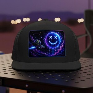 Può includere: Cappellino da camionista nero con una grafica al neon di una faccina sorridente e un UFO nello spazio. Il design utilizza colori viola e blu, creando un tema cosmico. Il cappello è su un tavolo nero, con un paesaggio desertico sullo sfondo.
