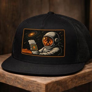 May include: Black trucker hat with a rectangular patch featuring an astronaut looking at a device, with a bigfoot silhouette in the helmet's visor. The background shows a galaxy and a red planet.