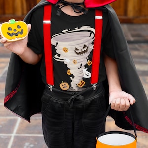 May include: A child dressed in a Halloween costume, wearing a black t-shirt with a cartoon tornado design, red suspenders, and a black cape. The child is holding a pumpkin-shaped cookie and an orange trick-or-treat bucket.