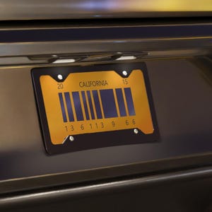 May include: A close-up of a car's rear end, showcasing a California license plate with a gold background and blue vertical lines. The plate has the text "CALIFORNIA" and numbers. The car is dark gray with dual exhaust pipes.