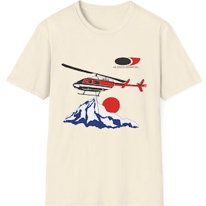 May include: Cream-colored t-shirt featuring a graphic of a red and black helicopter, a mountain range, and a red sun. The text "AIR SERVICES INTERNATIONAL" is above the helicopter. The design is printed on a short-sleeved tee.